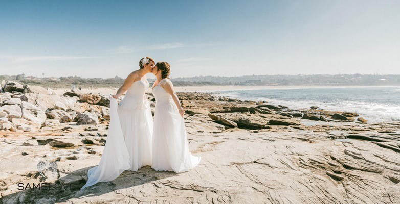 two brides kissing on rocks near the beach